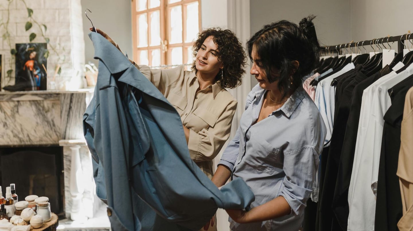 Two individuals working in a store are looking at a piece of clothing in a retail store. They stand near a clothing rack and a table with various items.